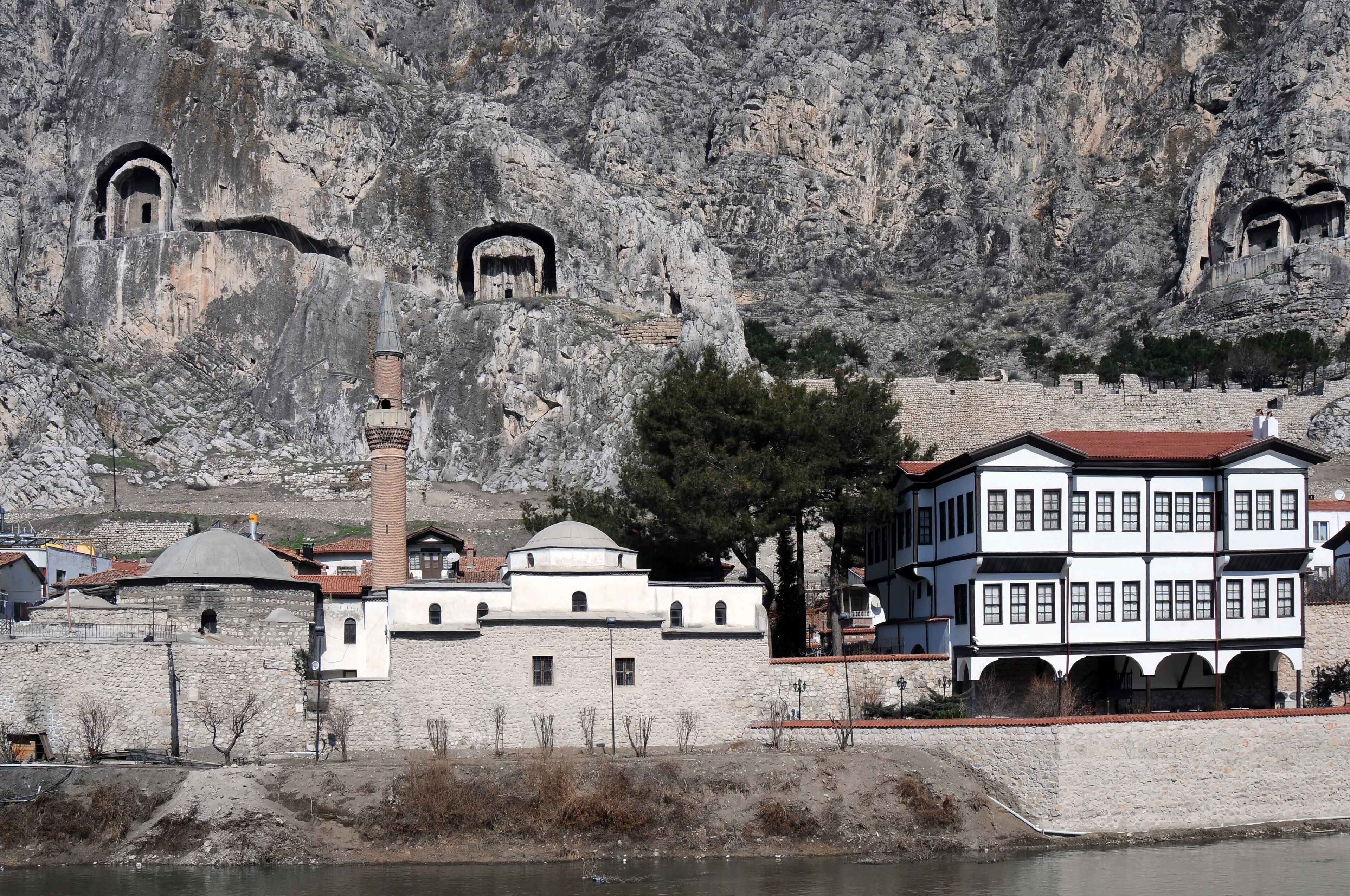 A historic stone building with a minaret, set against a rocky hillside with ancient rock-cut tombs.