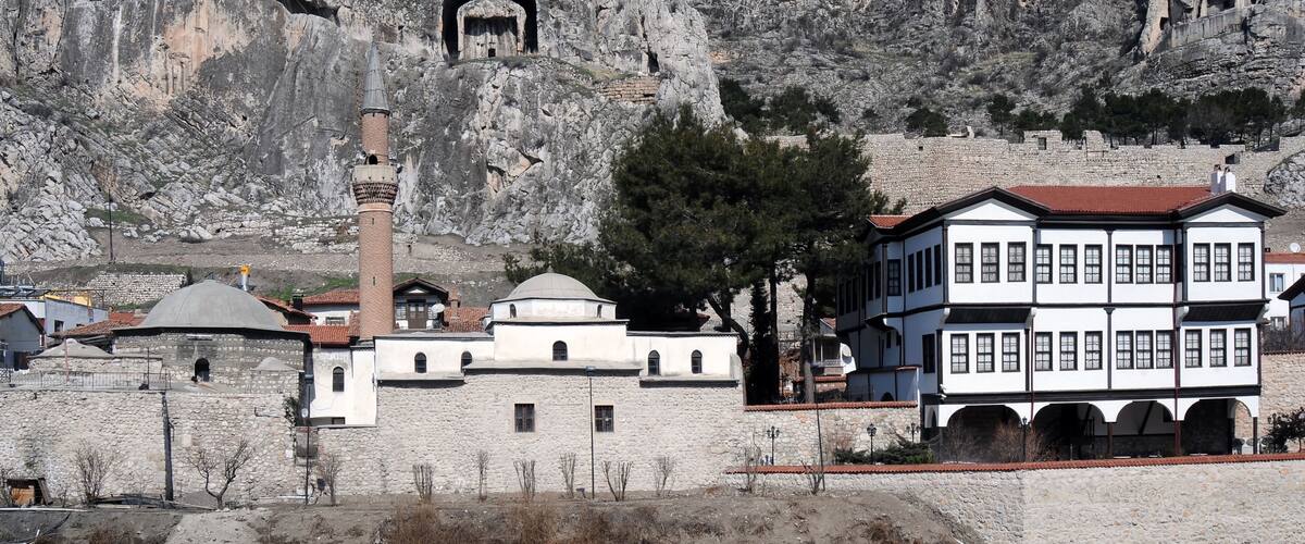 A historic stone building with a minaret, set against a rocky hillside with ancient rock-cut tombs.