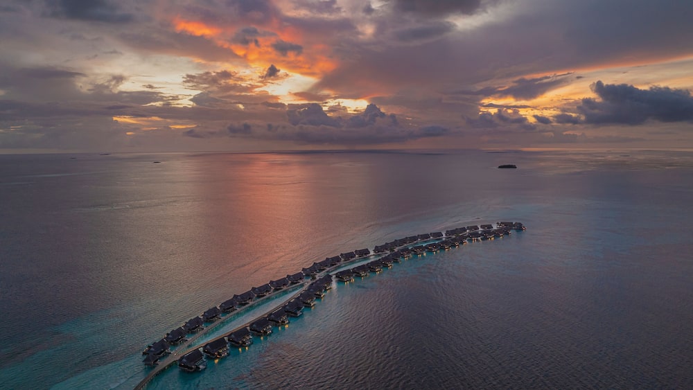 Une vue aérienne d’une longue structure incurvée s’étendant au-dessus de l’eau, avec des bâtiments reliés entre eux, le tout entouré d’eaux océaniques calmes et surplombé par un ciel spectaculaire au coucher du soleil.