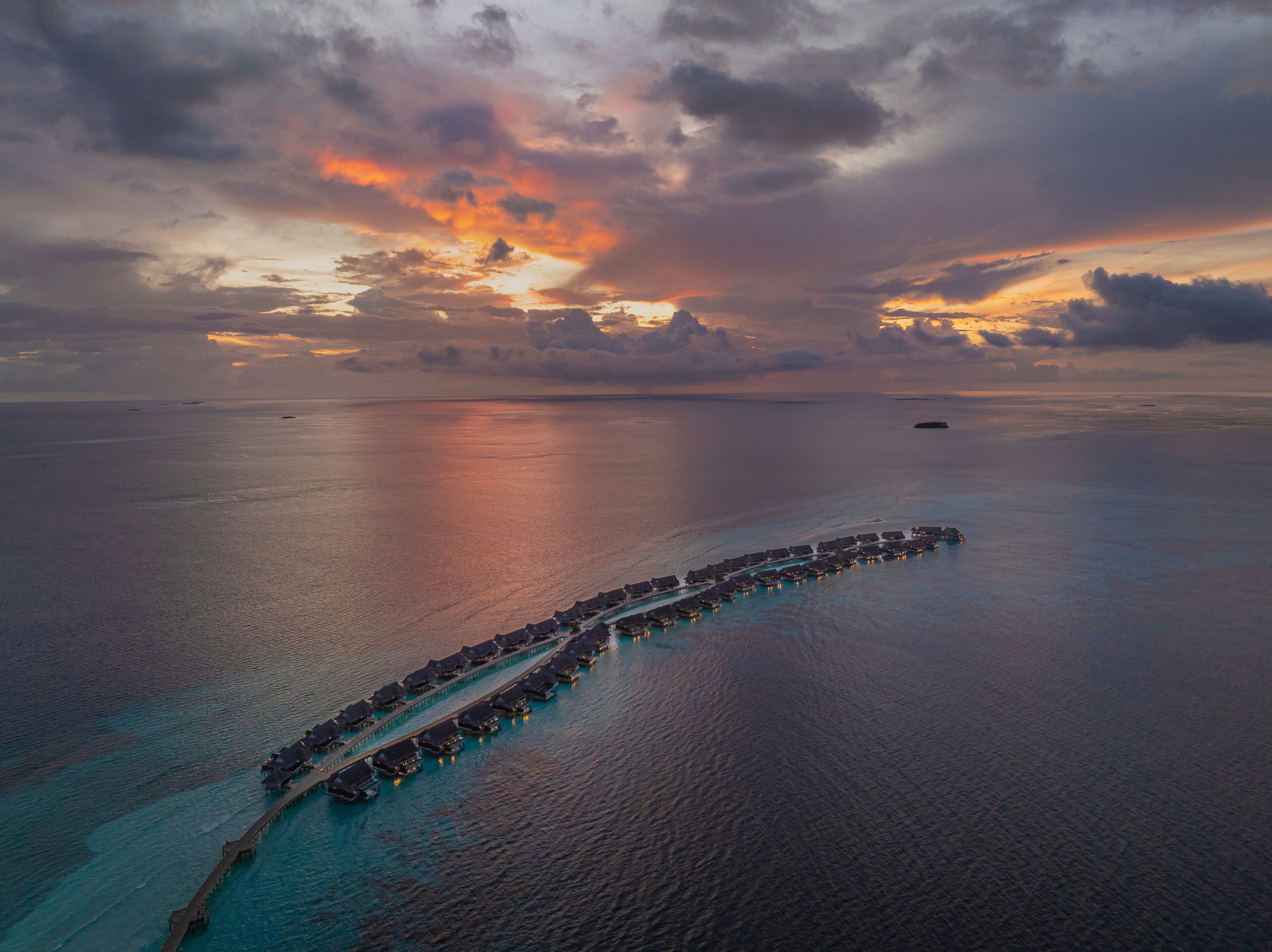 An aerial view of a long, curved overwater structure with connected buildings, surrounded by calm ocean waters and a dramatic sunset sky.