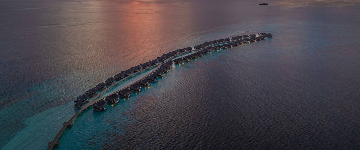 An aerial view of a long, curved overwater structure with connected buildings, surrounded by calm ocean waters and a dramatic sunset sky.
