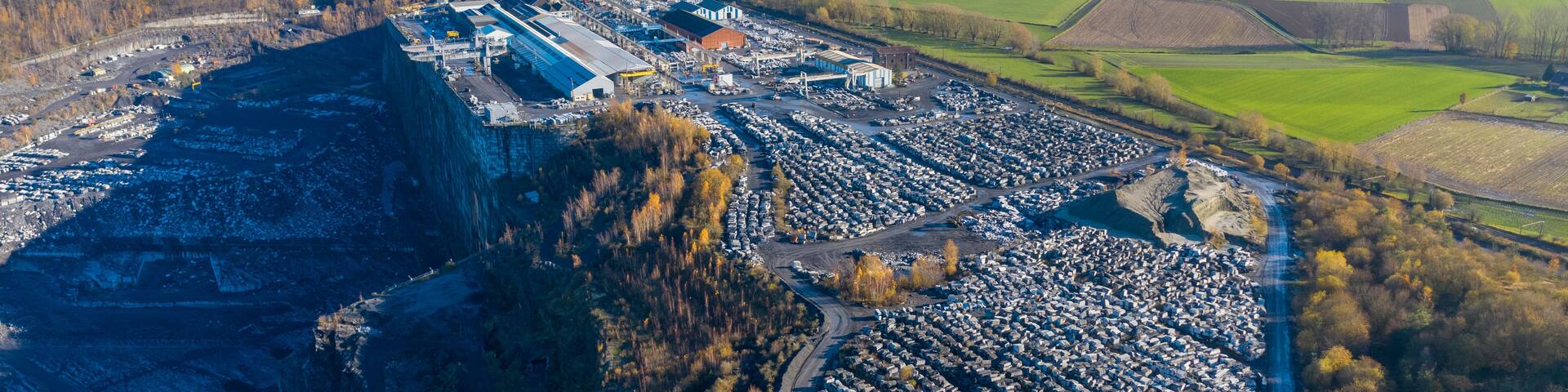 An aerial view of a quarry with large piles of rocks and a nearby industrial area with buildings and parked vehicles.