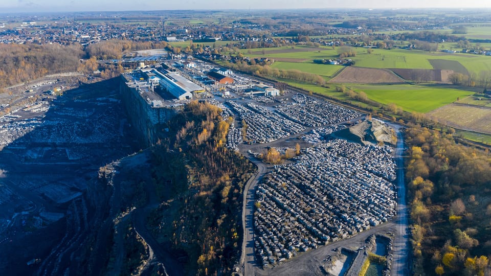 Vista aérea de una cantera con grandes montones de rocas y una zona industrial cercana, con edificios y vehículos estacionados.