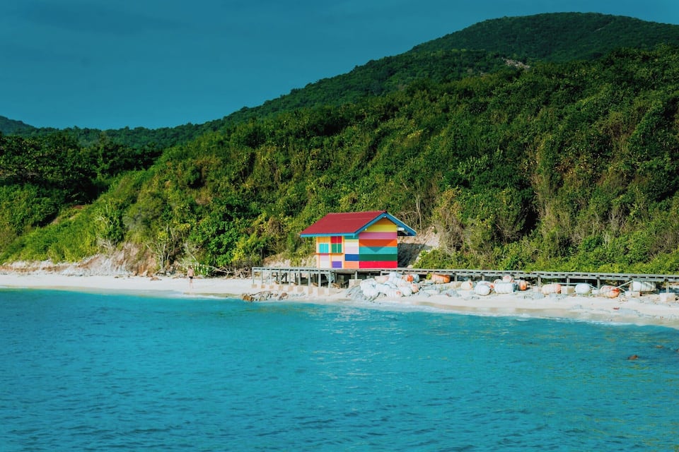 A colorful house with a red roof on a beach, surrounded by a forested hillside and clear blue waters.