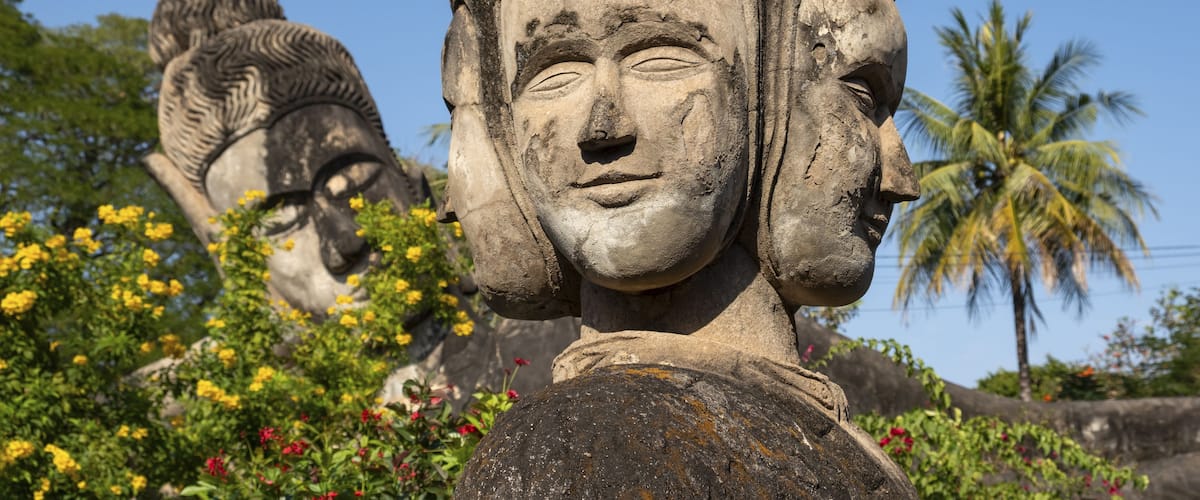 A large stone Buddha statue with intricate carvings, surrounded by lush greenery and other statues.