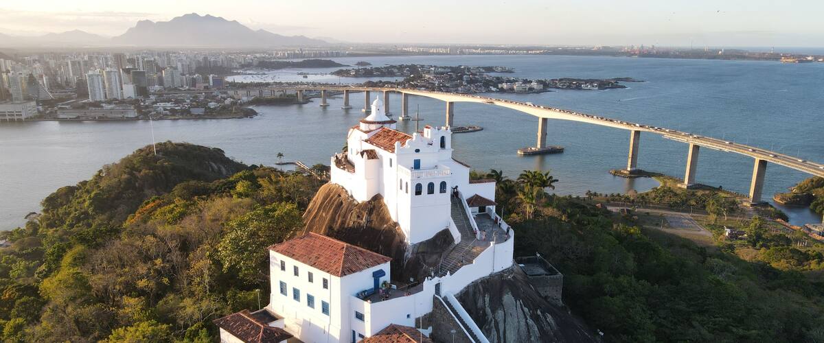 Igreja branca com telhado vermelho numa colina com vista para um corpo de água, uma ponte e uma paisagem urbana ao longe.