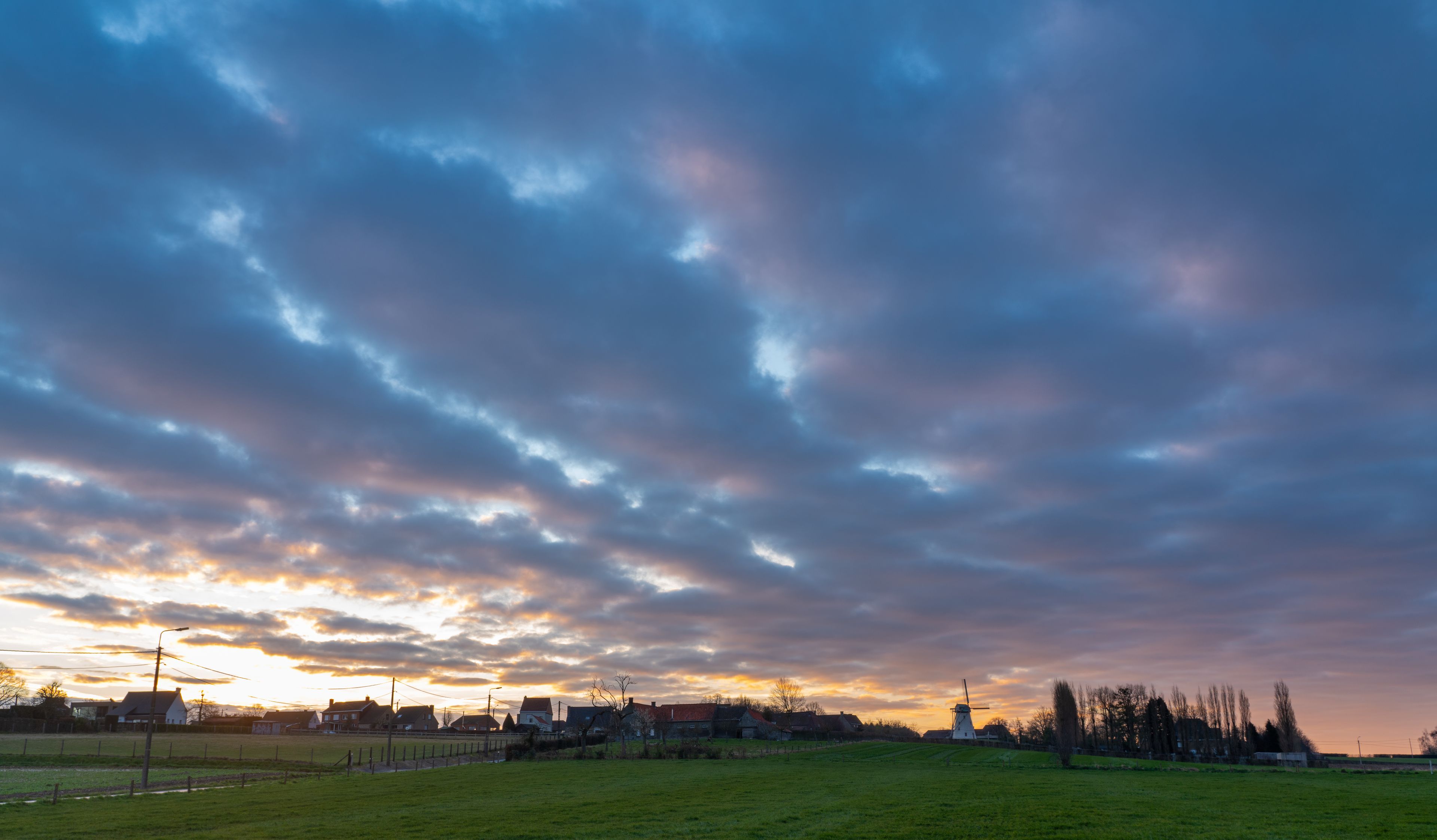 Een landelijk landschap met een windmolen, huizen en een bewolkte lucht bij zonsondergang.