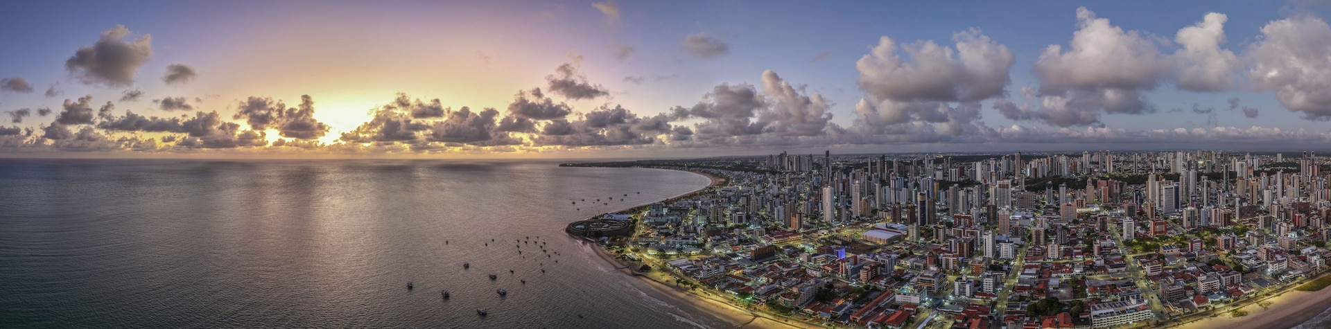 Paisagem urbana costeira com uma linha de costa sinuosa, edifícios urbanos densos e um céu ao pôr do sol.