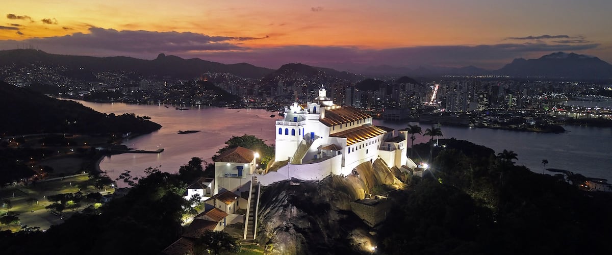 A coastal cityscape at dusk with a prominent white church on a hill, a river, and a city skyline in the background.