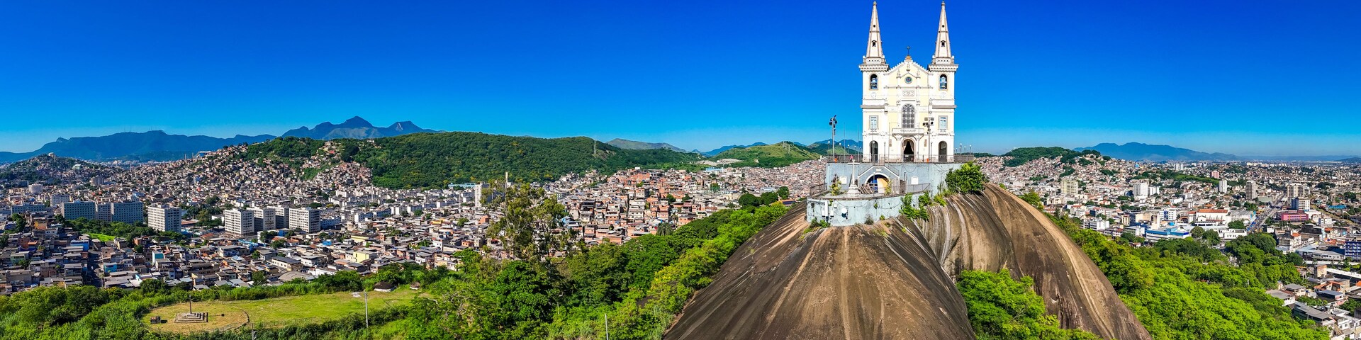 Uma catedral com duas torres, cercada por uma paisagem urbana densamente povoada e colinas verdes ao fundo.