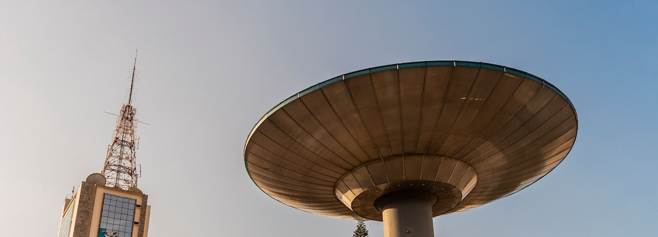 Uma grande estrutura de antena parabólica, um edifício alto com antena visível e uma estrada com semáforos e veículos.