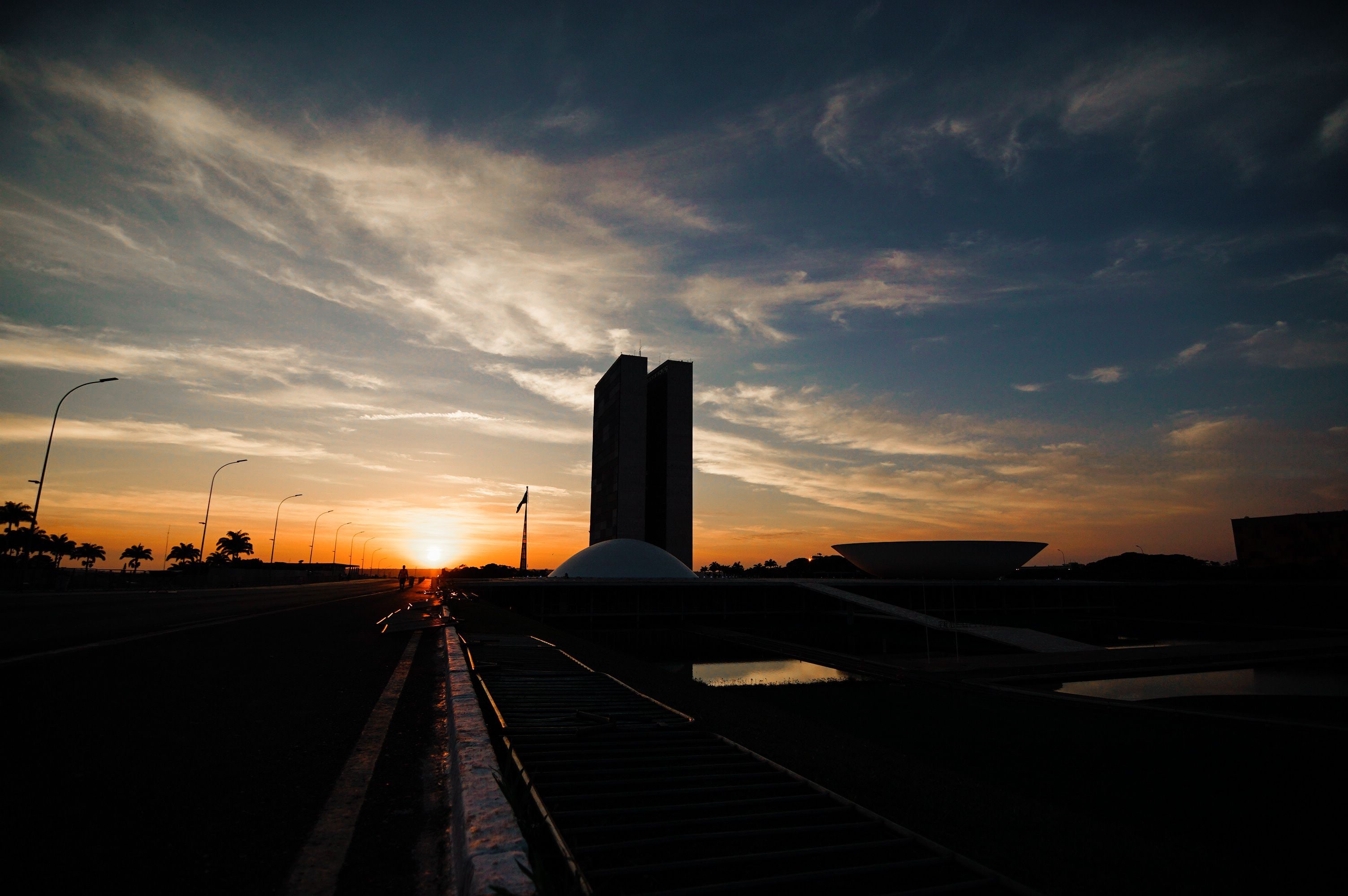 Um pôr do sol sobre uma ponte, com um prédio alto e palmeiras ao fundo.