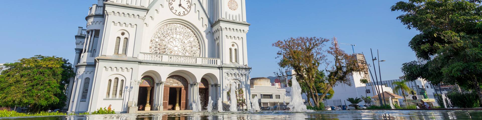 A white cathedral with a large clock and three spires, reflecting in a body of water.