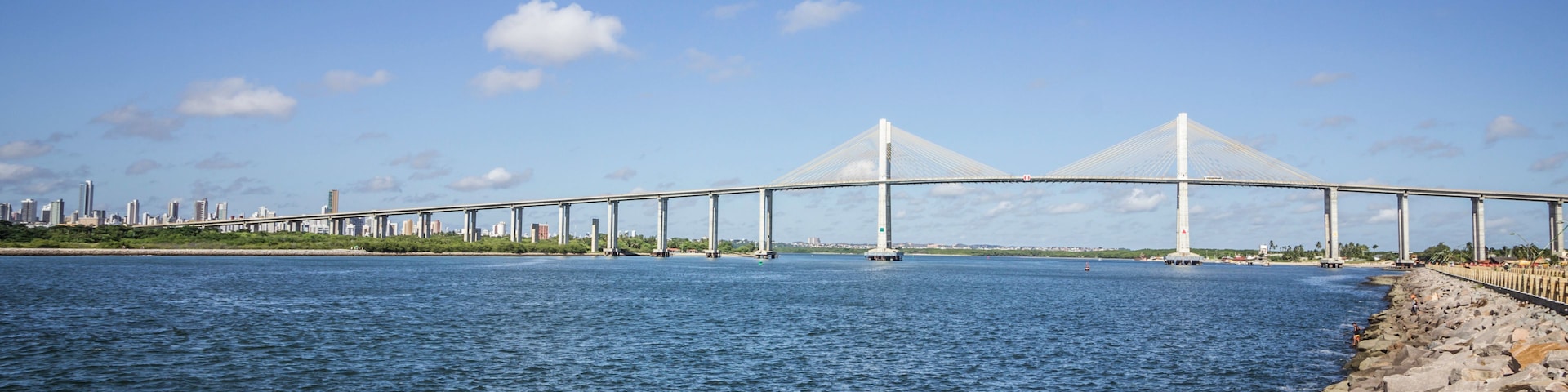 Un puente colgante imponente que cruza un río ancho, con el horizonte de una ciudad al fondo.