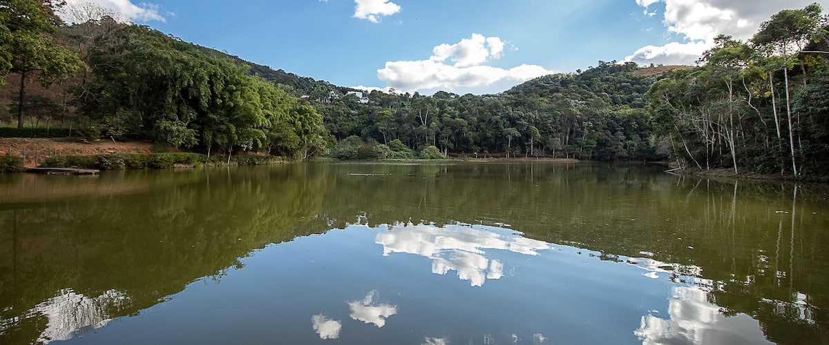 Um lago tranquilo cercado por floresta densa e colinas sob um céu parcialmente nublado.