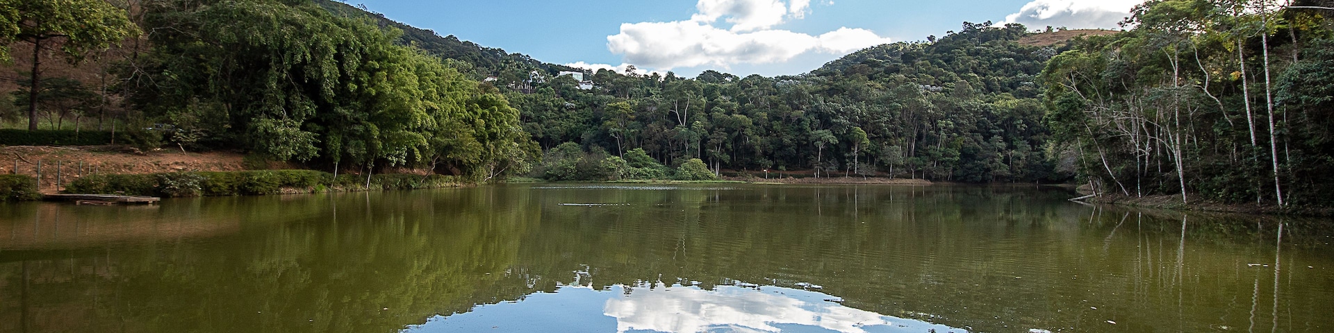 Um lago tranquilo cercado por floresta densa e colinas sob um céu parcialmente nublado.