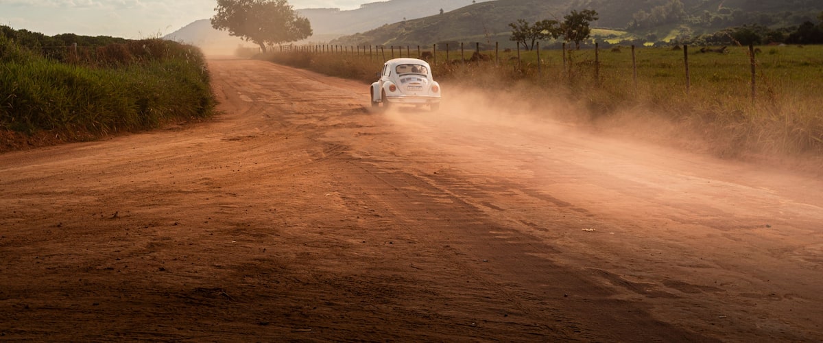 A white car driving on a dusty road with a fence on one side and a tree on the other.