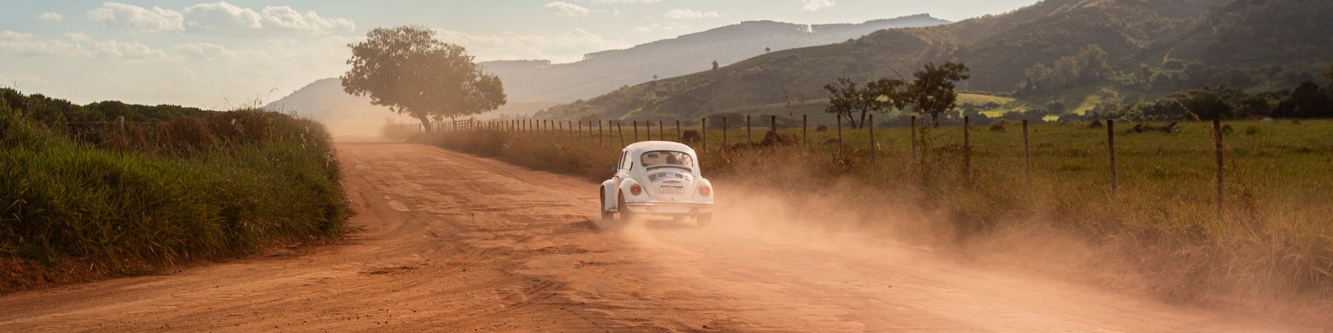 Um carro branco dirigindo em uma estrada de terra, com uma cerca de um lado e uma árvore do outro.
