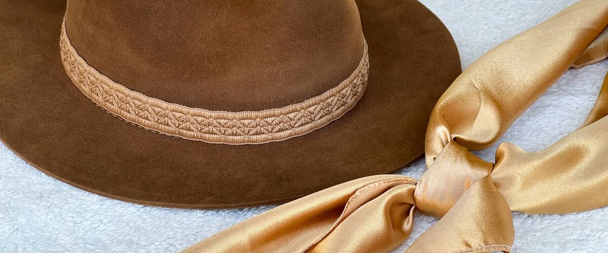 A brown hat with a braided band and a gold ribbon tied around it.
