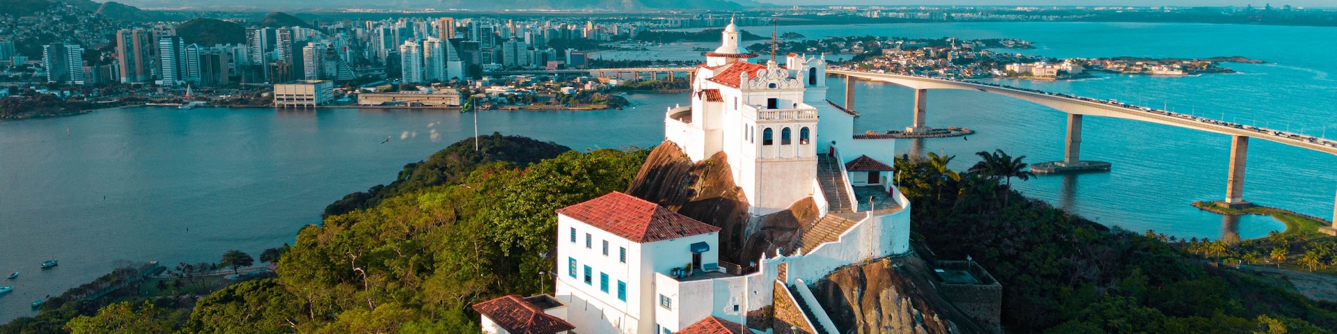 Paisagem costeira com uma estrutura imponente em estilo de castelo numa colina, uma ponte sobre a água e uma área urbana densa ao fundo.