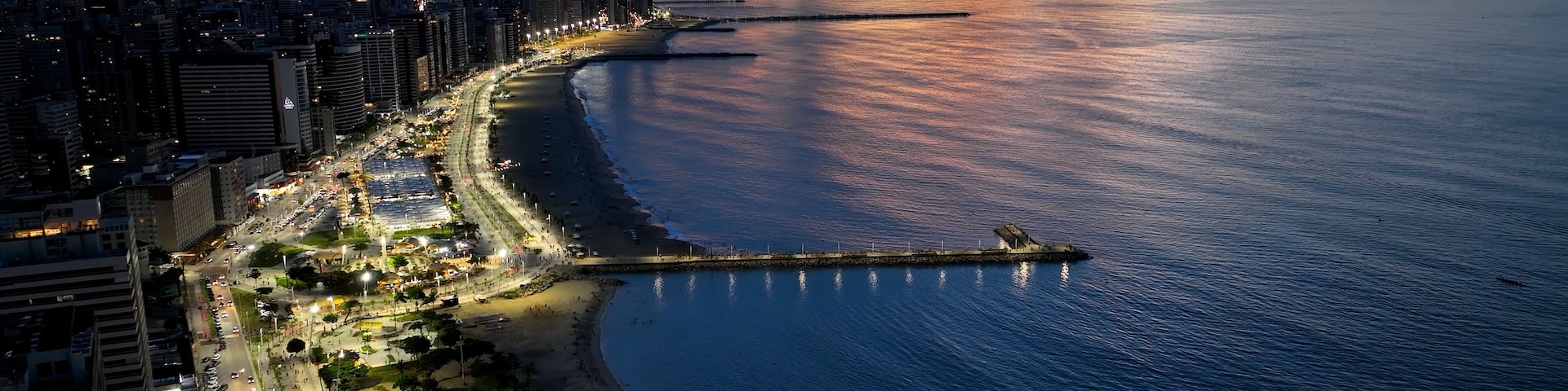 Paisagem costeira ao pôr do sol, com um cais imponente que se estende para o oceano, área urbana densamente povoada com arranha-céus e uma orla marítima bem iluminada.