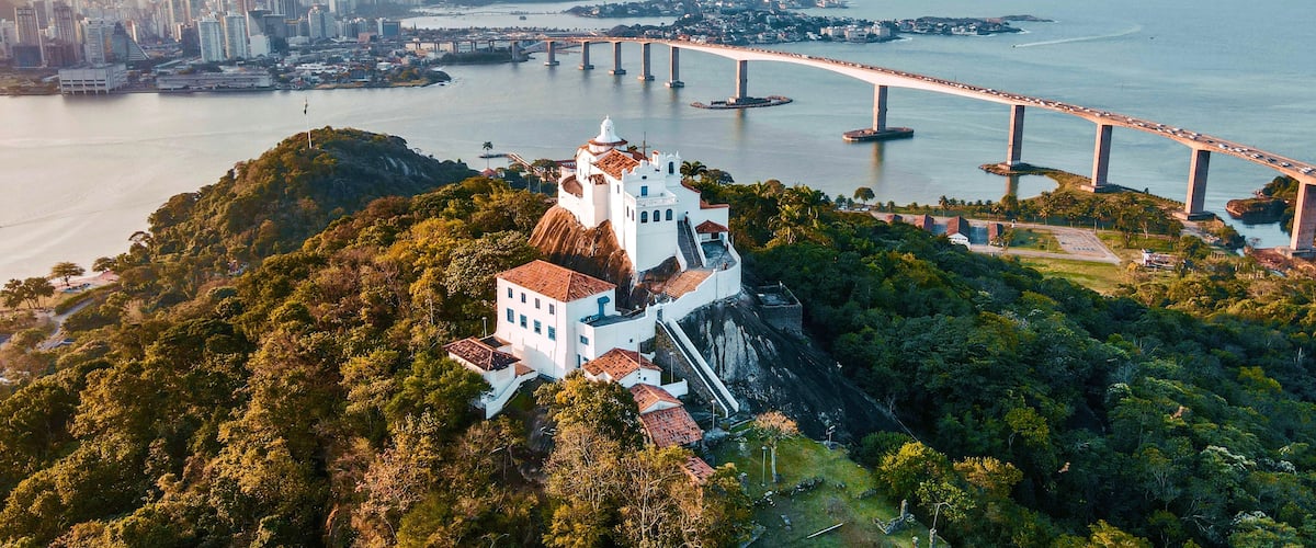 Paisagem costeira urbana com uma ponte notável, vegetação exuberante e um edifício histórico branco com telhado vermelho.