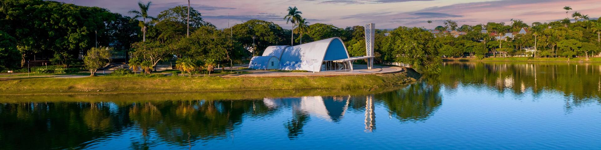 A serene lake with a modern building featuring a blue roof, surrounded by lush greenery and palm trees.