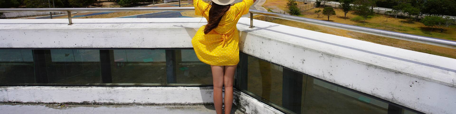 A person in a yellow dress with arms raised, overlooking a coastal cityscape with buildings and a clear blue sky.