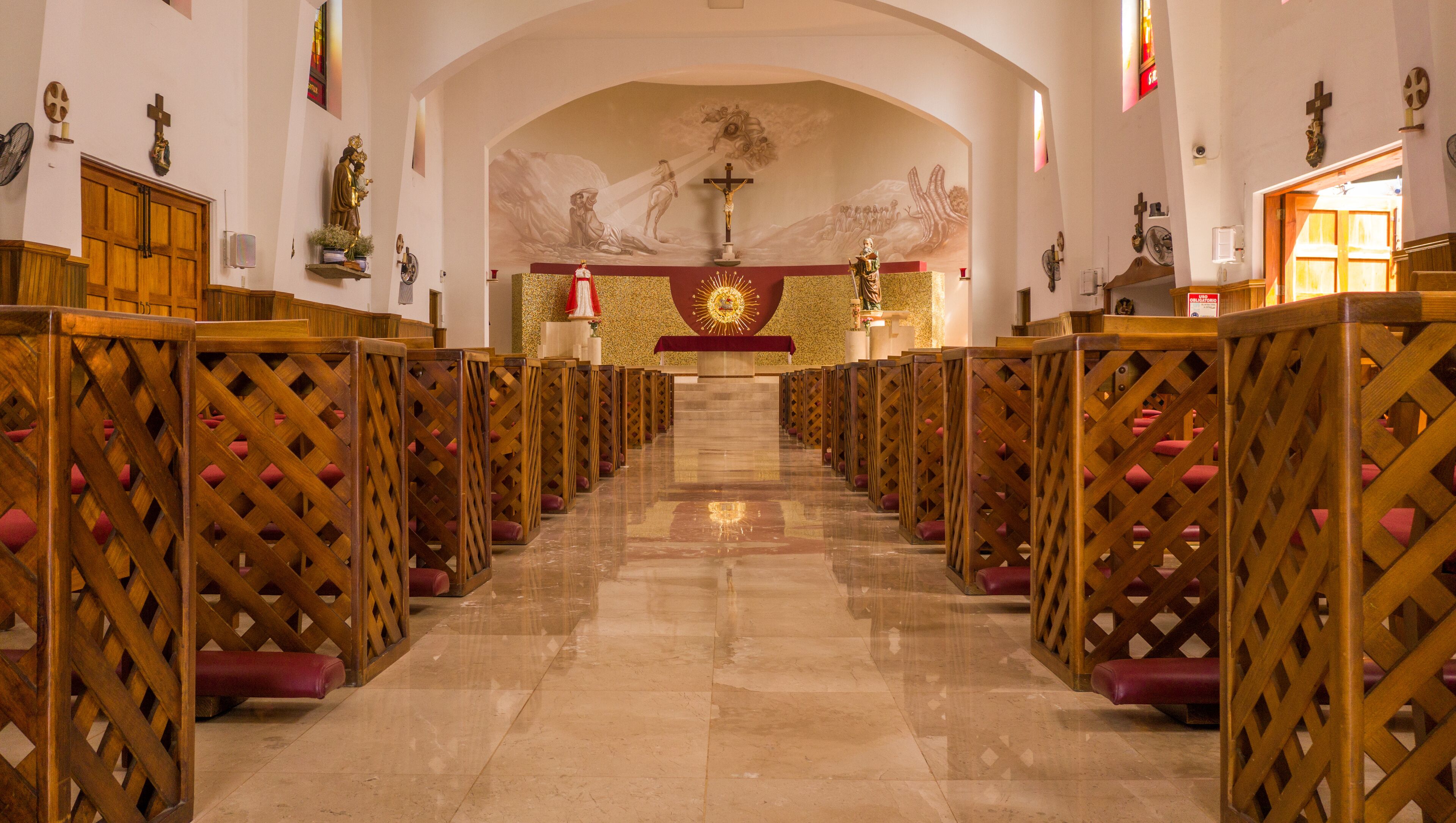 A church interior with rows of wooden pews, a central altar with a crucifix, and religious statues on the walls.