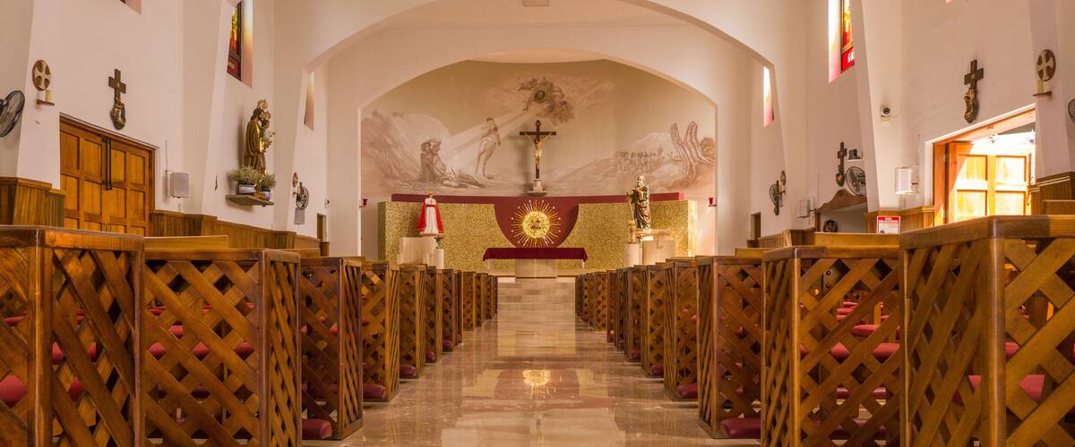 A church interior with rows of wooden pews, a central altar with a crucifix, and religious statues on the walls.