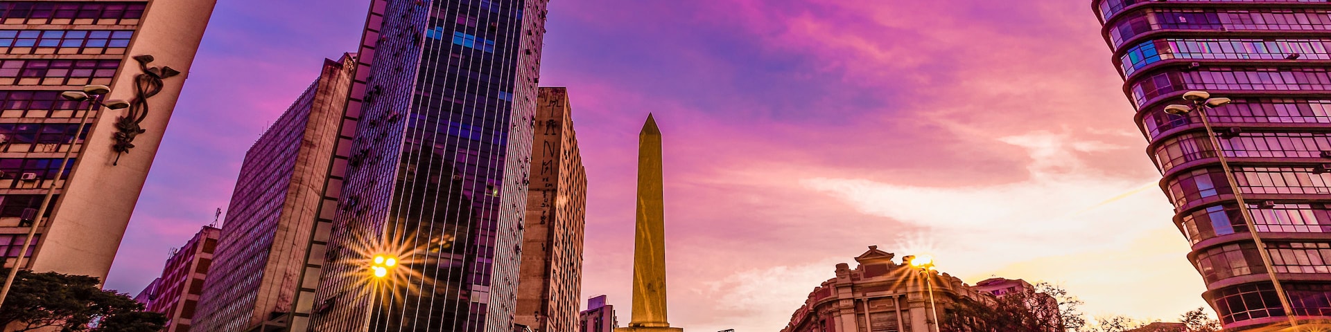 Paisagem urbana com edifícios altos, um monumento e uma rua com rastros de luz.