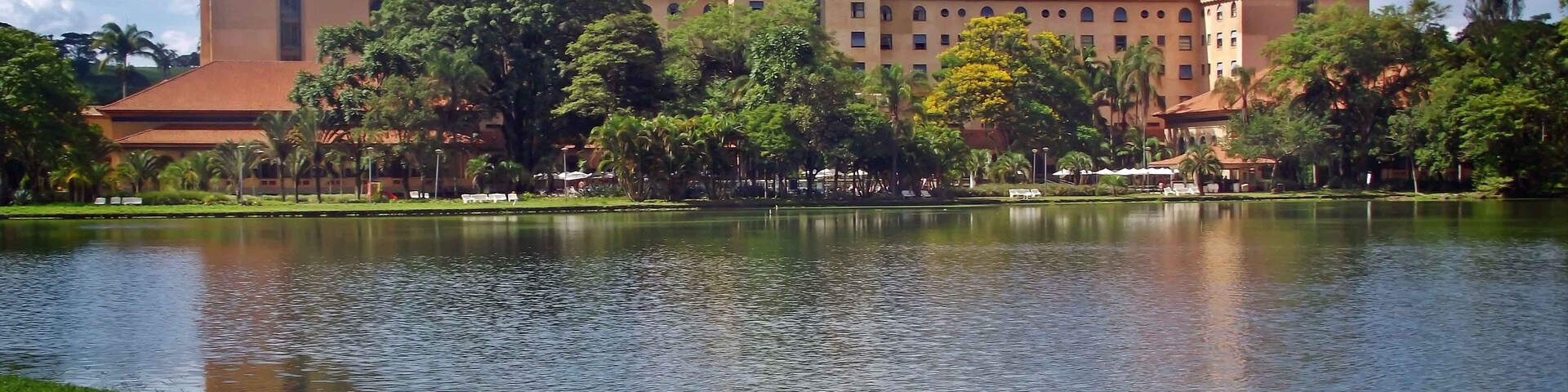 Un edificio grande con techo rojo y ventanas arqueadas, rodeado de árboles y un lago tranquilo.