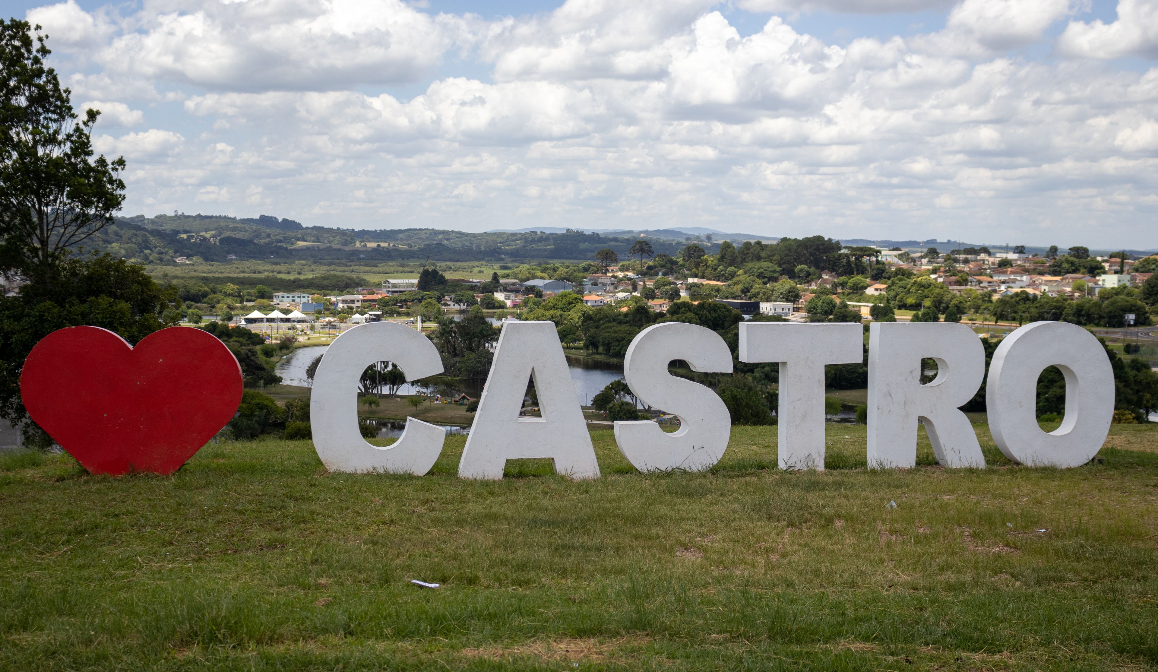 Un letrero con la palabra 'CASTRO' y un símbolo de corazón, frente a un paisaje de un pueblo y colinas.