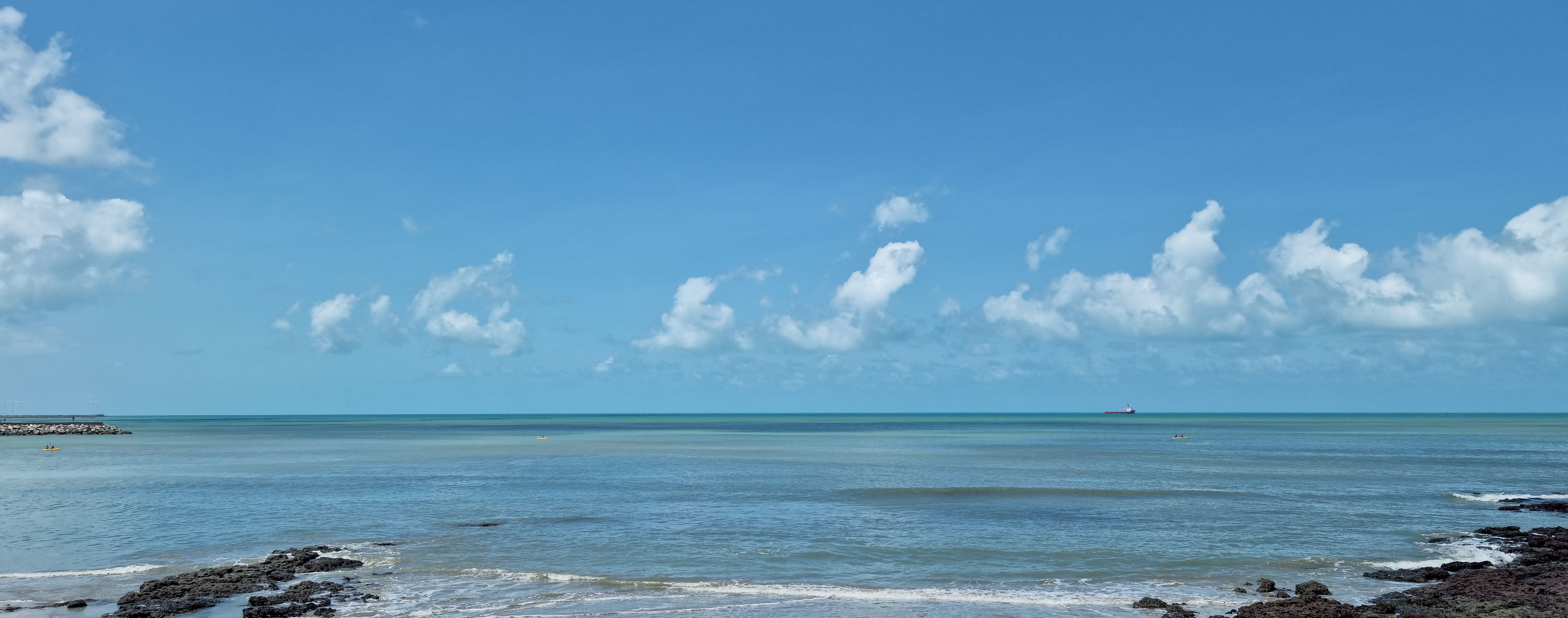 Escena costera con cielos despejados, mar tranquilo, costa rocosa y barcos a lo lejos.