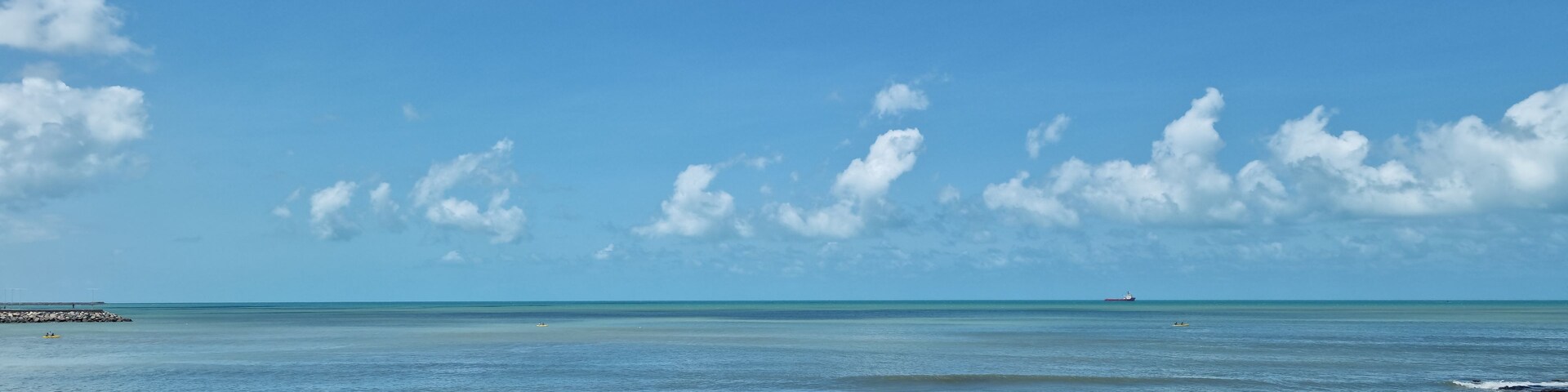 Escena costera con cielos despejados, mar tranquilo, costa rocosa y barcos a lo lejos.