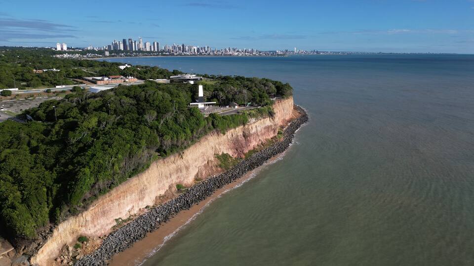 A coastal cityscape with a prominent cliff, dense greenery, and a calm sea.