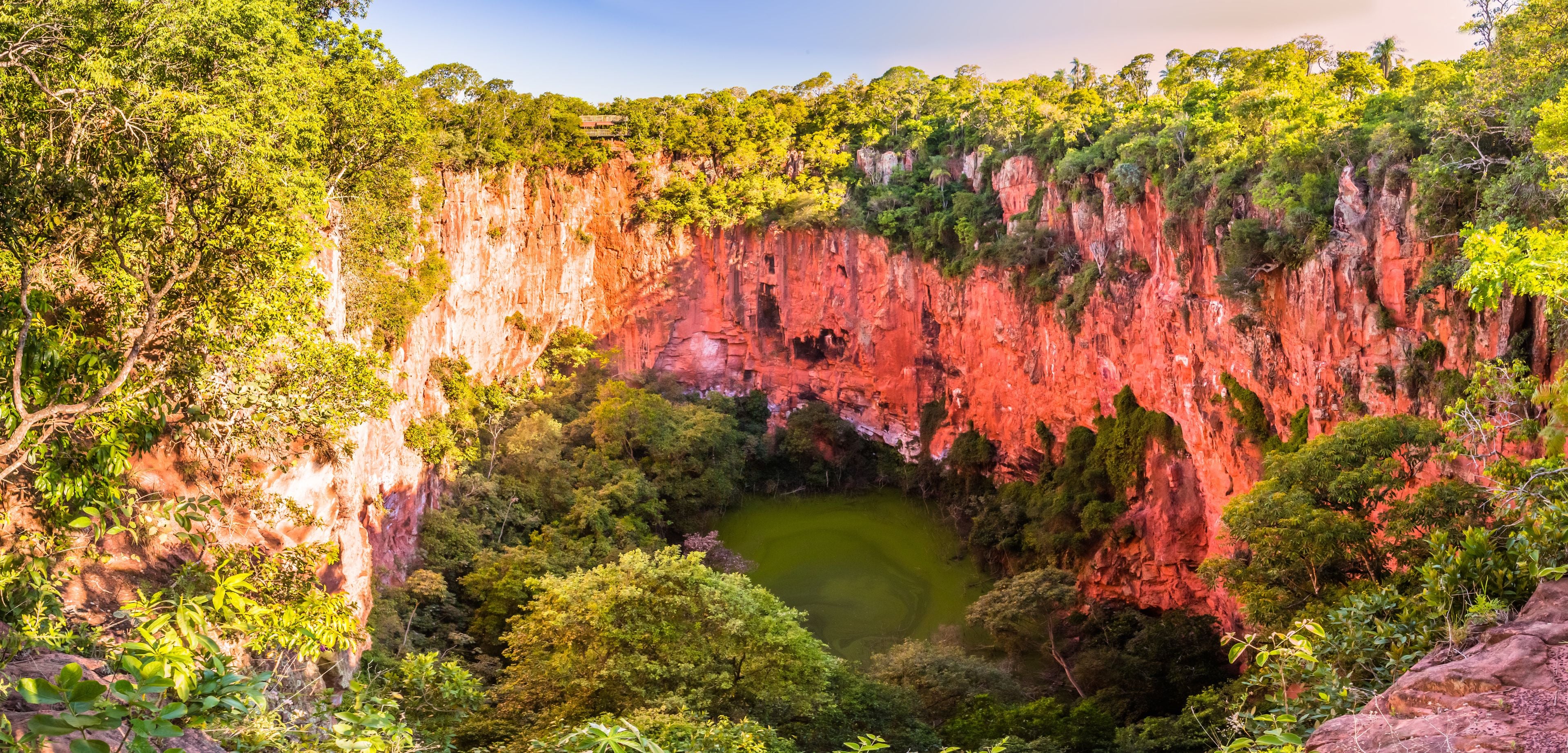 Um desfiladeiro profundo com penhascos vermelhos íngremes e um pequeno lago verde cercado por vegetação densa.