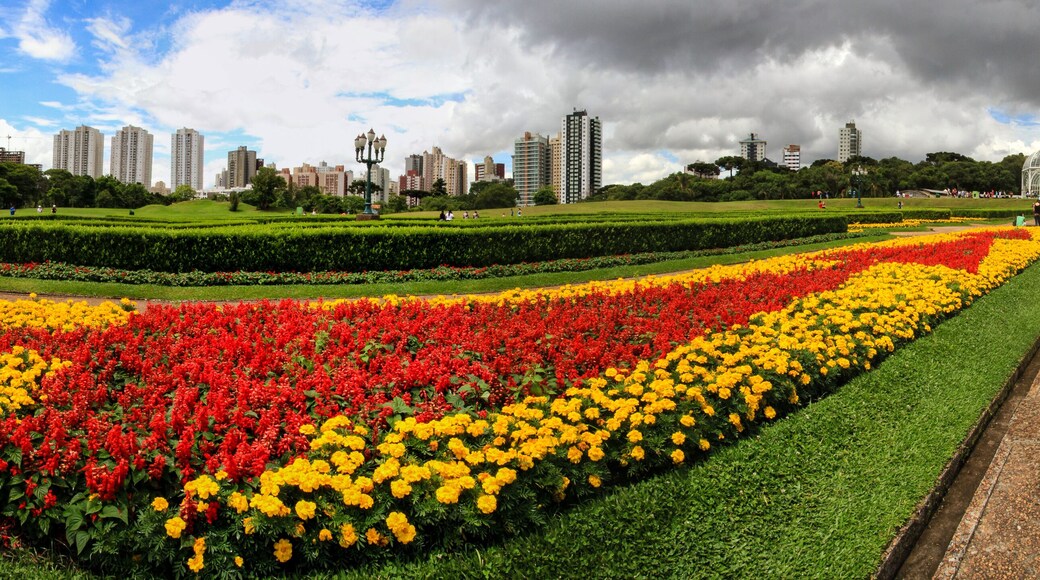 Panorama Botanischer Garten in Curitiba Brasilien mit roten, gelben und grünen Pflanzen und Blumen, gläsernem Gewächshaus und Stadtpanorama am Horizont