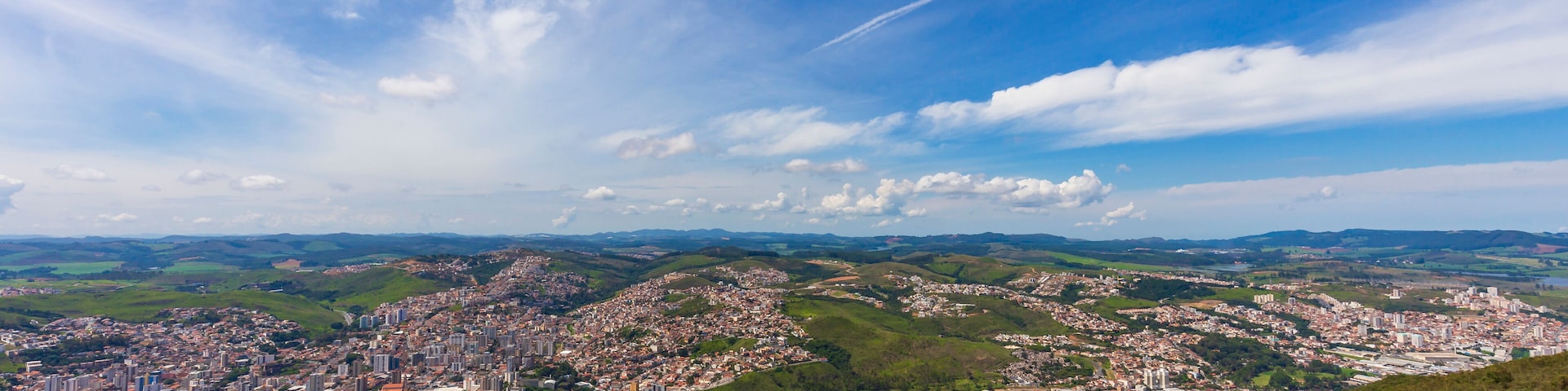Vista panorâmica de uma cidade com áreas urbanas e verdes, céu claro e colinas ao longe.
