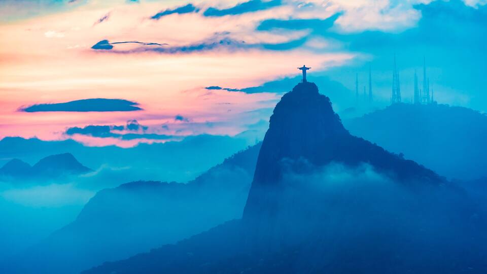 A mountain peak with a statue at the top, surrounded by clouds and other mountains.