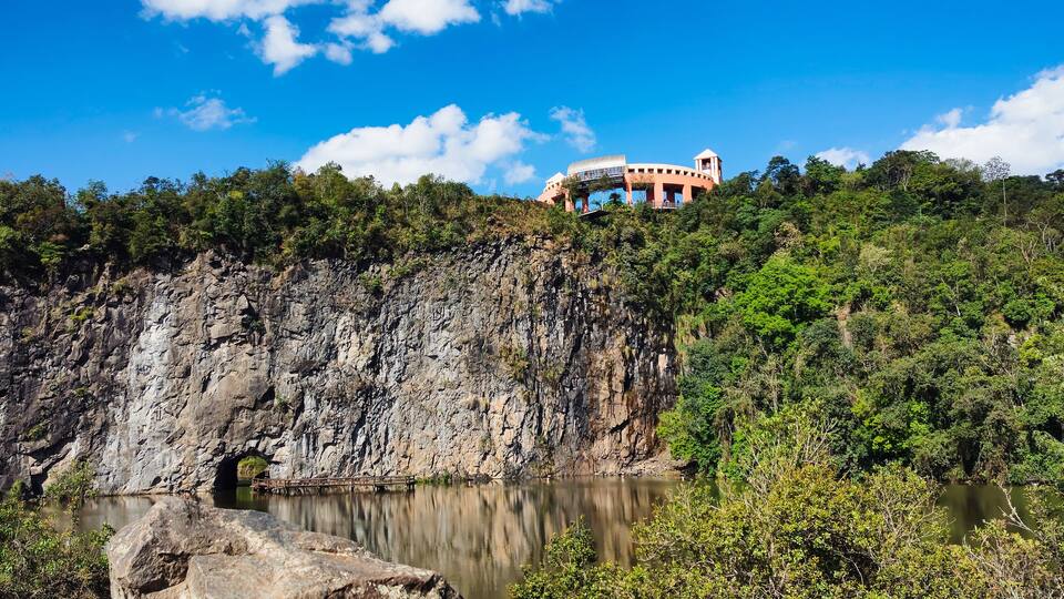 A rocky cliff with a cave entrance, a lake, and a building on top of the cliff.