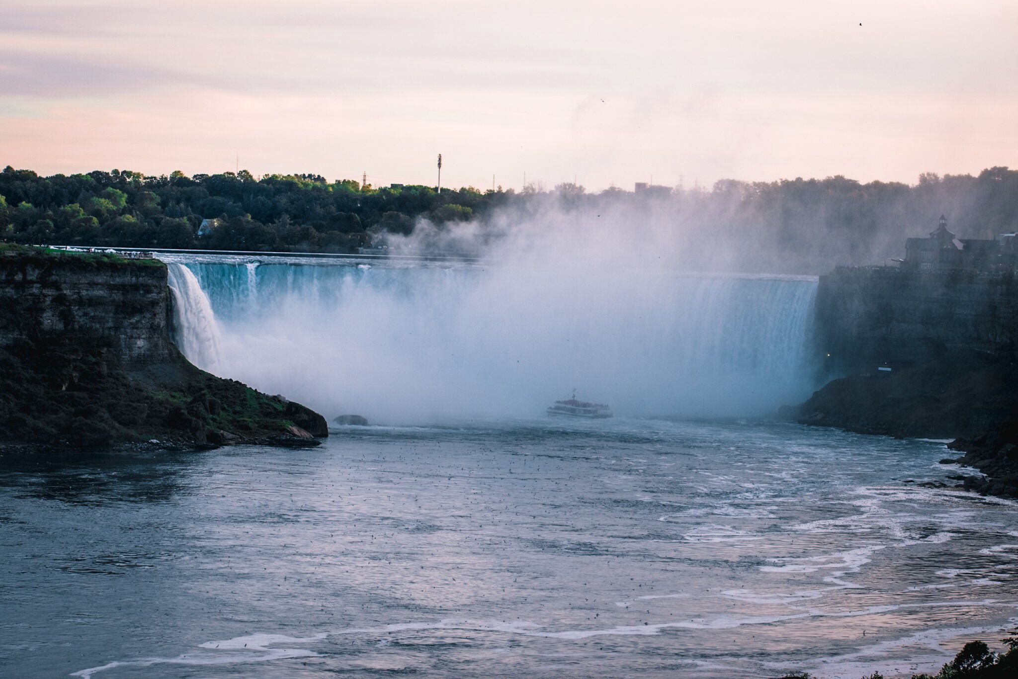 A large waterfall with mist rising from the base, surrounded by rocky cliffs and a distant city skyline.
