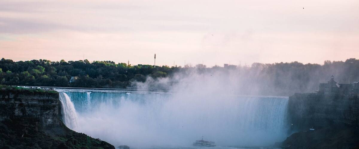 A large waterfall with mist rising from the base, surrounded by rocky cliffs and a distant city skyline.