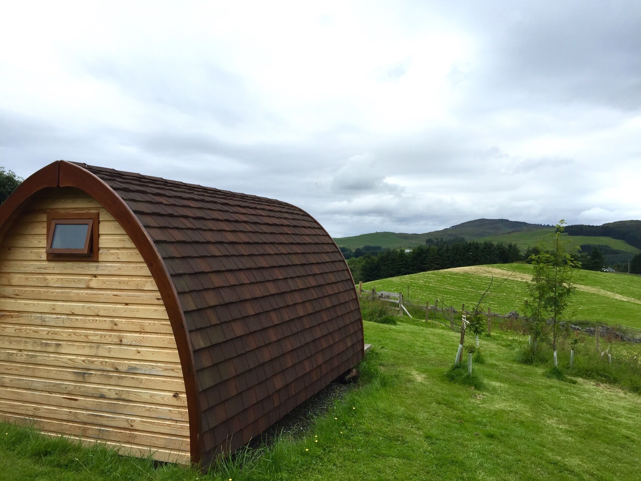 A wooden house with a sloped roof in a grassy field with hills in the background.