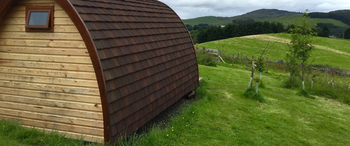 A wooden house with a sloped roof in a grassy field with hills in the background.