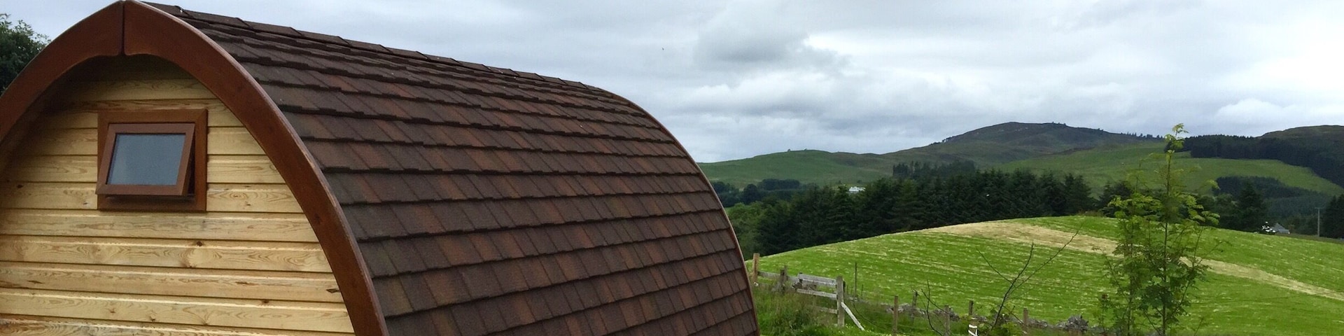 A wooden house with a sloped roof in a grassy field with hills in the background.