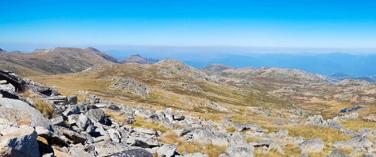 A mountainous landscape with rocky terrain and a distant view of layered mountain ranges under a clear blue sky.