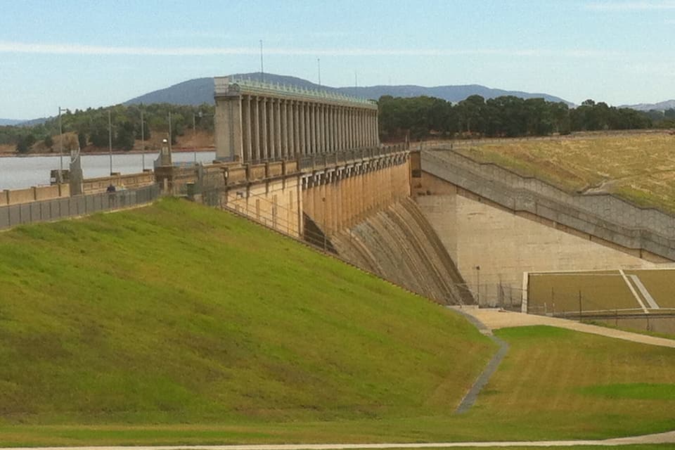 Un barrage avec un grand réservoir en amont, entouré de collines et bordé par une étendue d’eau.