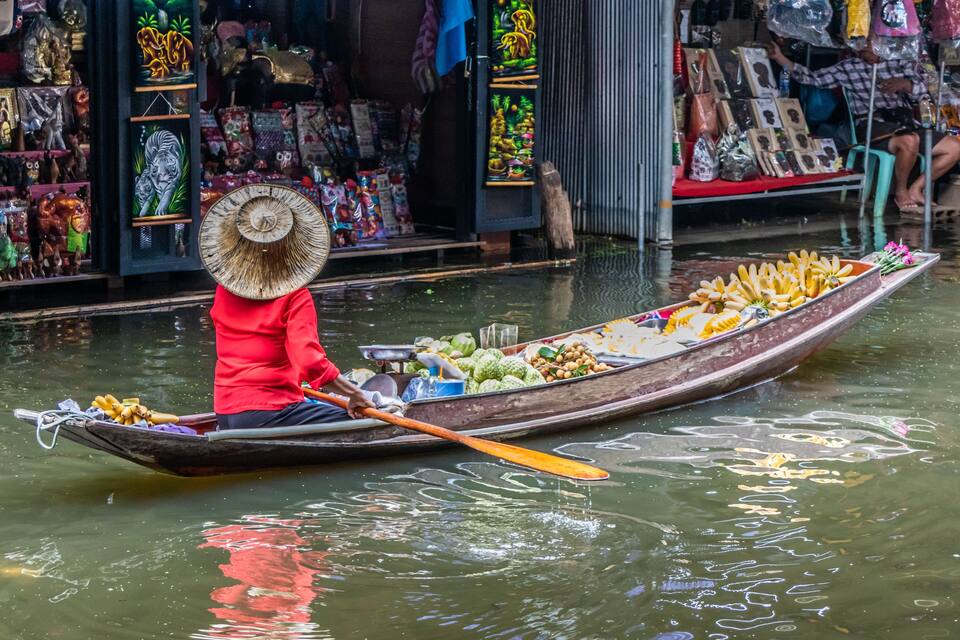 Eine Person in roter Kleidung und Strohhut rudert in einem langen Boot voller Erntegüter und Waren.