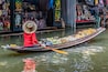 Eine Person in roter Kleidung und Strohhut rudert in einem langen Boot voller Erntegüter und Waren.